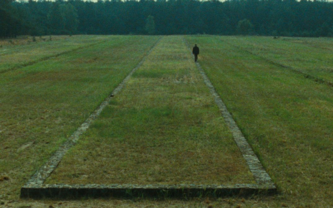 Filmstill aus SHOAH: Zu sehen ist eine Landschaft mit Wiese, im Hintergrund Wald. Auf der Wiese sind die Überreste einer Bebauung zu erkennen, eine Person geht daran entlang.