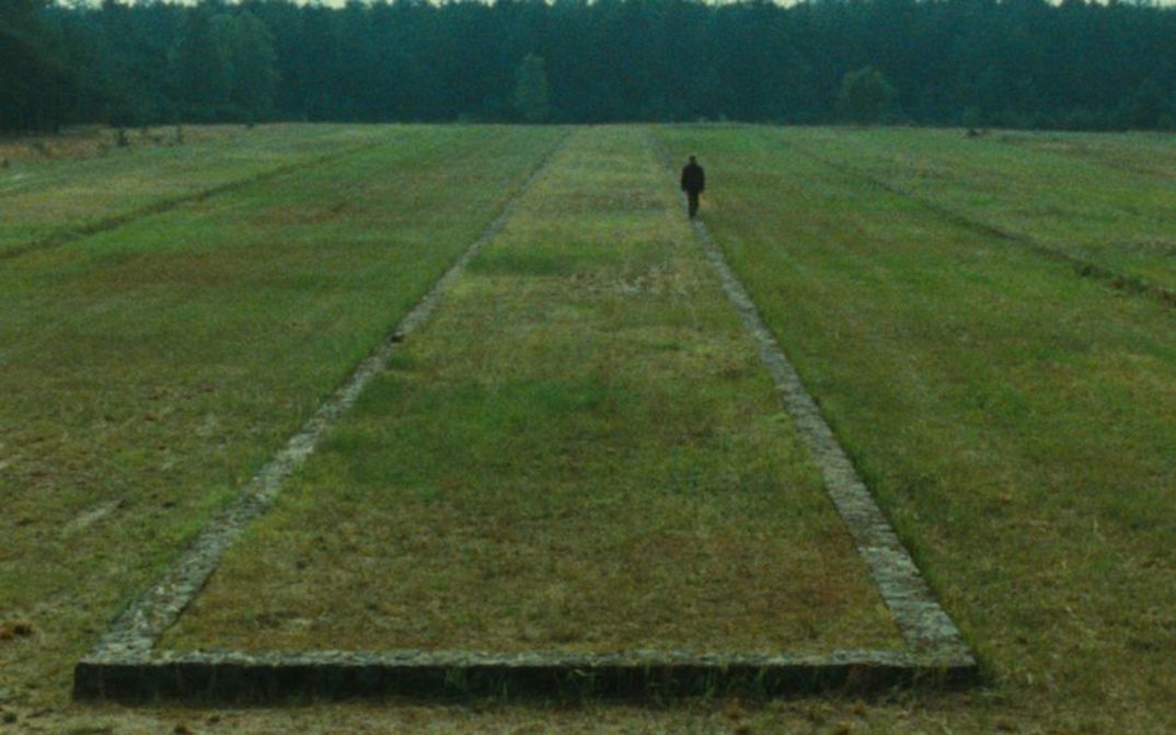 Film still from SHOAH: You can see a landscape with a meadow and forest in the background. The remains of a building can be seen in the meadow, a person is walking along it.