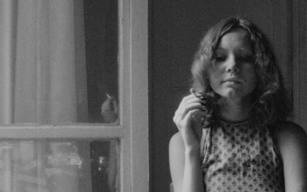 Film still from LE 15/8: A young woman with long, curly hair is standing by the window in an apartment, holding a cigarette between her fingers.