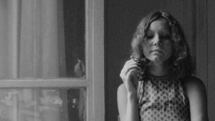 Film still from LE 15/8: A young woman with long, curly hair is standing by the window in an apartment, holding a cigarette between her fingers.