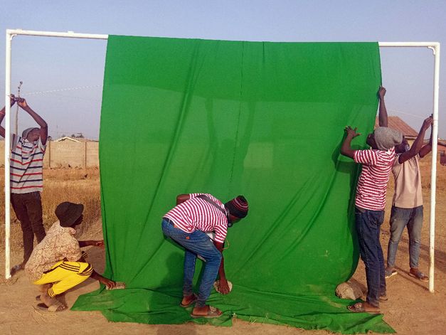 Mehrere Personen spannen im Freien ein großes grünes Tuch an einem Gestell, Steine beschweren den Stoff am Boden.