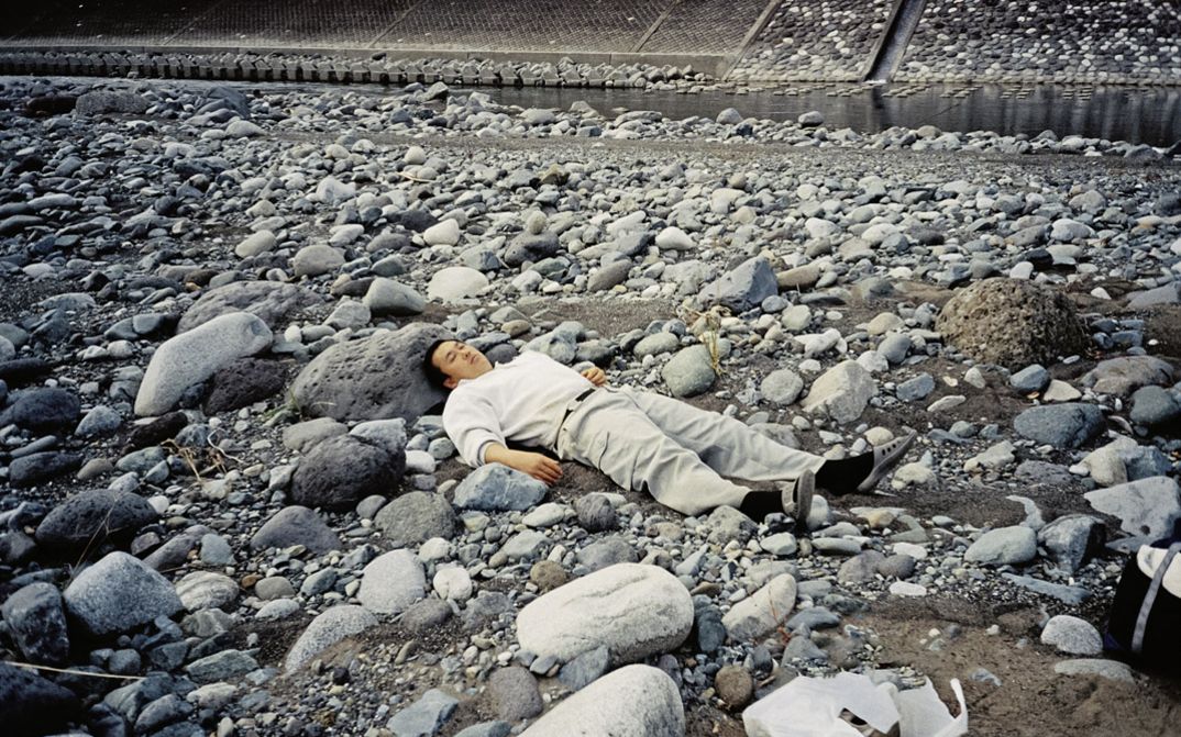 Filmstill aus dem Film "Ishi ga aru" von Tatsunari Ota. Ein Mann in heller Kleidung liegt auf grauen Steinen. Im Hintergrund fließt zwischen den Steinen ein Bach. 