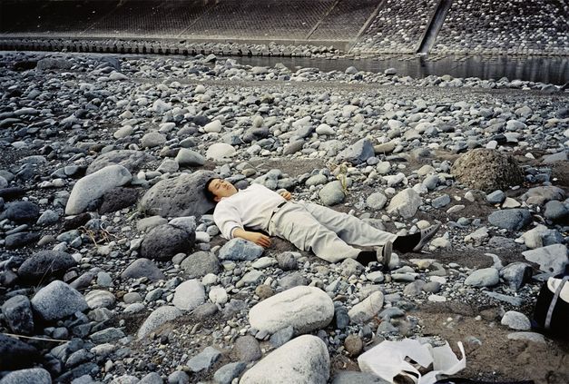Still from the film "Ishi ga aru" by Tatsunari Ota. A man in light clothing is lying on top of grey stones. There is a small stream of water flowing behind the rocks in the background.