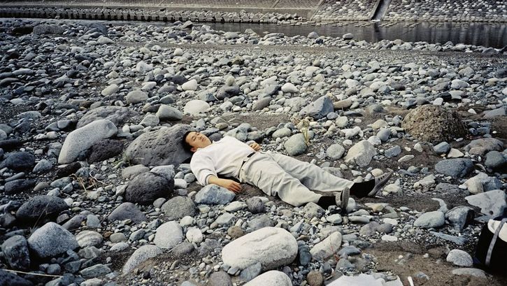 Still from the film "Ishi ga aru" by Tatsunari Ota. A man in light clothing is lying on top of grey stones. There is a small stream of water flowing behind the rocks in the background.