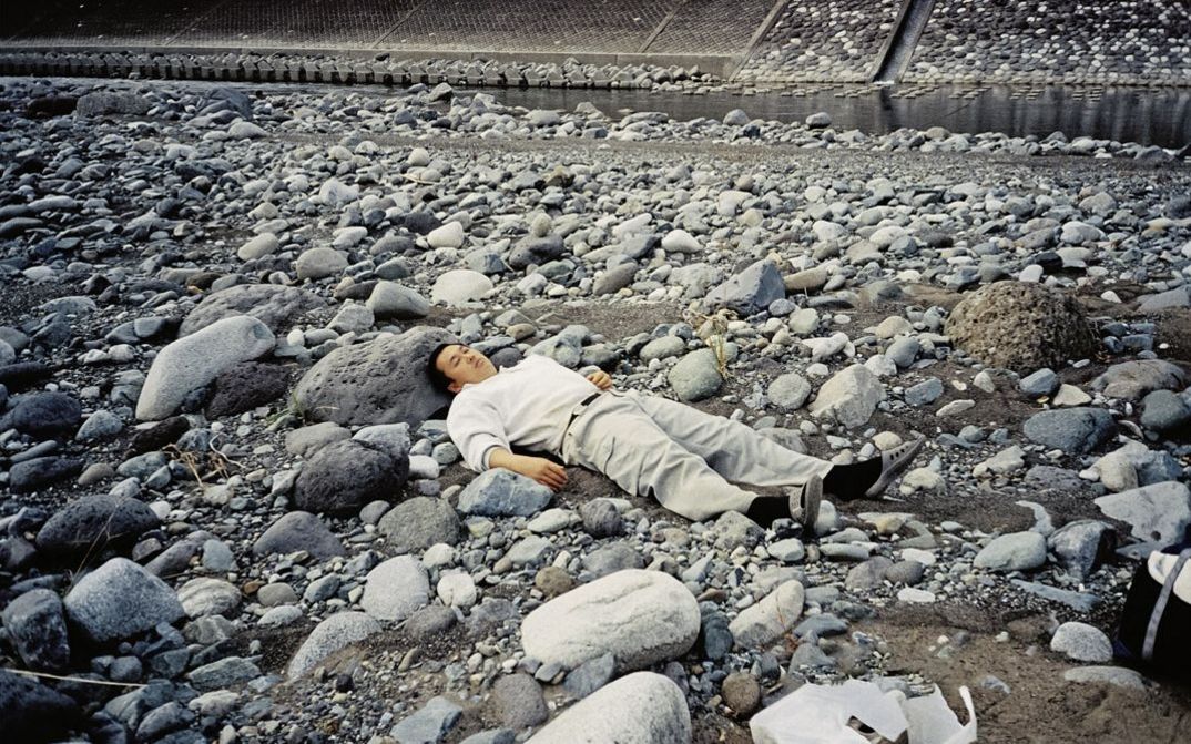 Still from the film "Ishi ga aru" by Tatsunari Ota. A man in light clothing is lying on top of grey stones. There is a small stream of water flowing behind the rocks in the background.