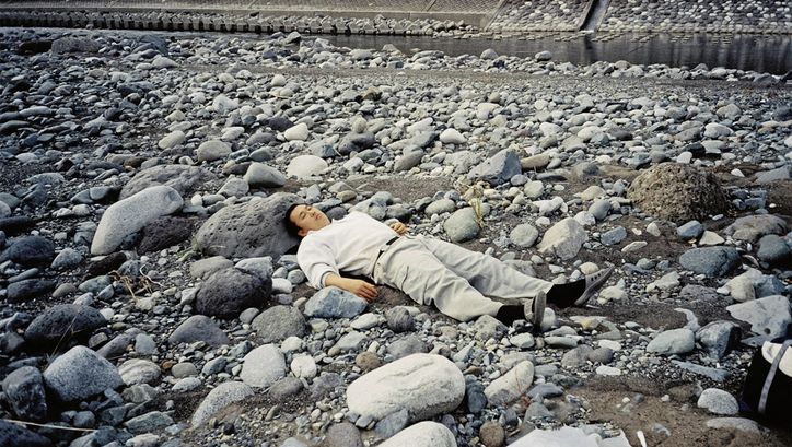 Filmstill aus dem Film "Ishi ga aru" von Tatsunari Ota. Ein Mann in heller Kleidung liegt auf grauen Steinen. Im Hintergrund fließt zwischen den Steinen ein Bach. 