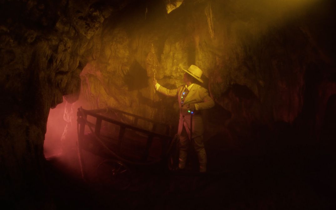 Film Still aus „Fanfictie: Volcanology“ von Riar Rizaldi. In einer Höhle steht eingetaucht in rötliches und gelbliches Licht eine Figur in pastellfarbenem Anzug und Hut.