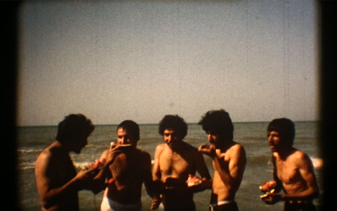 Still from the film "Majmouan" by Mohammadreza Farzad. Men standing in front of the sea eating watermelon.