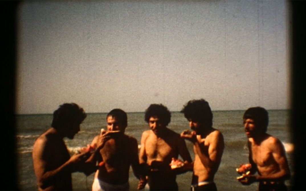 Filmstill aus dem Film "Majmouan" von Mohammadreza Farzad. Männer stehen vor dem Meer und essen Wassermelone.