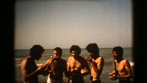 Still from the film "Majmouan" by Mohammadreza Farzad. Men standing in front of the sea eating watermelon.