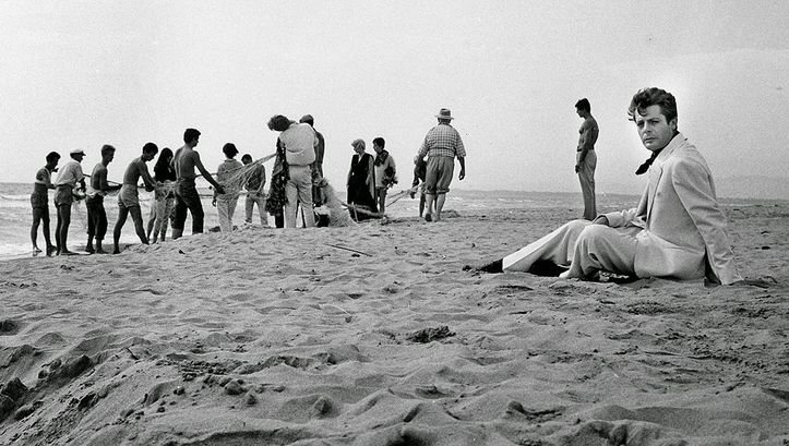 Film still from LA DOLCE VITA: Marcello Mastroianni sits in a suit on a beach, behind him fishermen are busy with a fishing net.