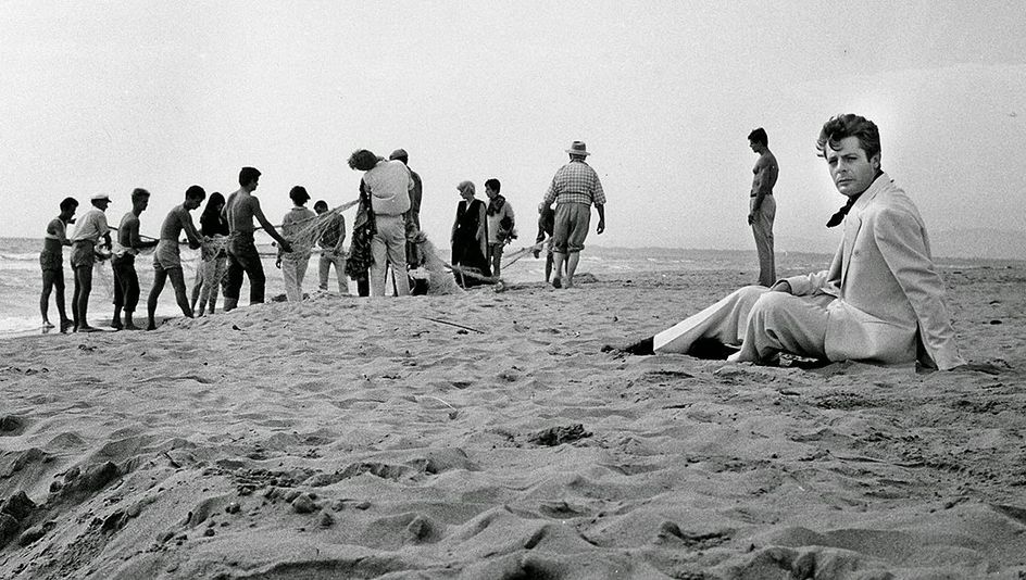Filmstill aus LA DOLCE VITA: Marcello Mastroianni sitzt im Anzug an einem Strand, hinter ihm sind Fischer mit einem Fischernetz beschäftigt.