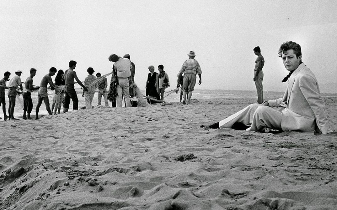 Filmstill aus LA DOLCE VITA: Marcello Mastroianni sitzt im Anzug an einem Strand, hinter ihm sind Fischer mit einem Fischernetz beschäftigt.