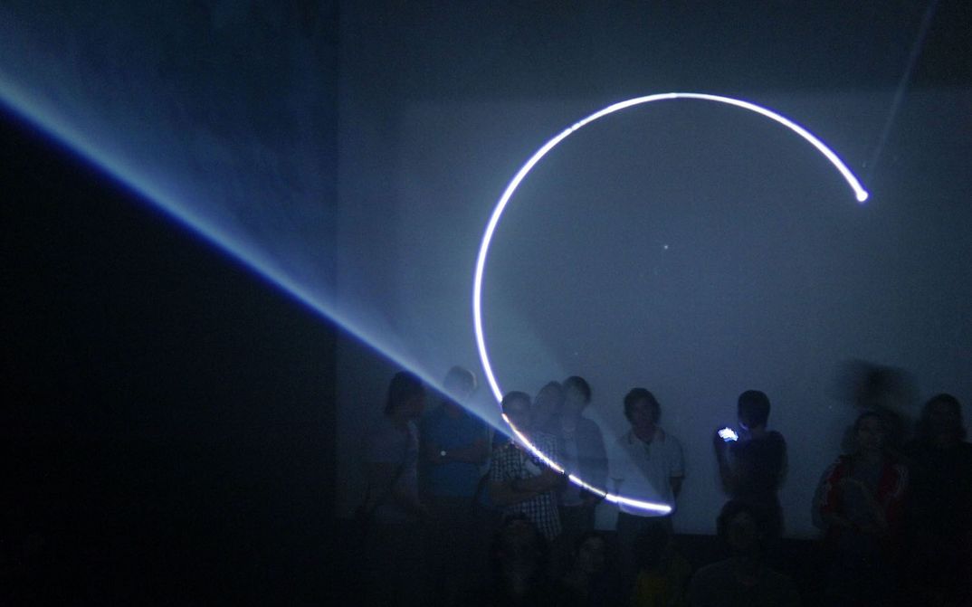Film still from LINE DESCRIBING A CONE: In a dark room, an almost closed circle is projected onto a screen. You can see the cone of light and several people standing in front of the screen, looking at the light.