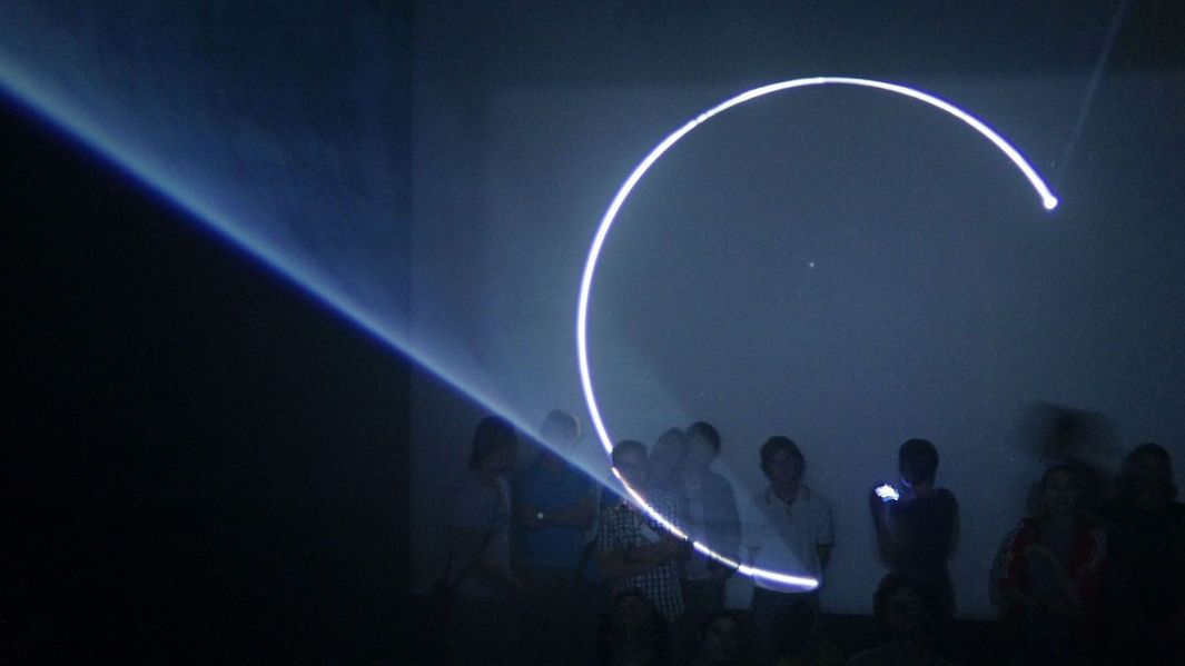 Filmstill aus LINE DESCRIBING A CONE: In einem dunklen Raum wird auf eine Leinwand ein fast geschlossener Kreis projiziert. Man sieht den Lichtkegel und einige Personen, die an der Leinwand stehen und das Licht betrachten.