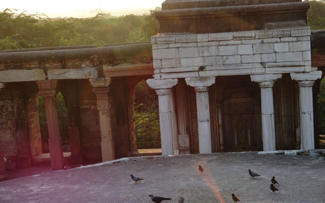 Film still from the film "No Stranger at All" by Priya Sen. A low sun shines on a temple ruin. Pigeons can be seen on the court.