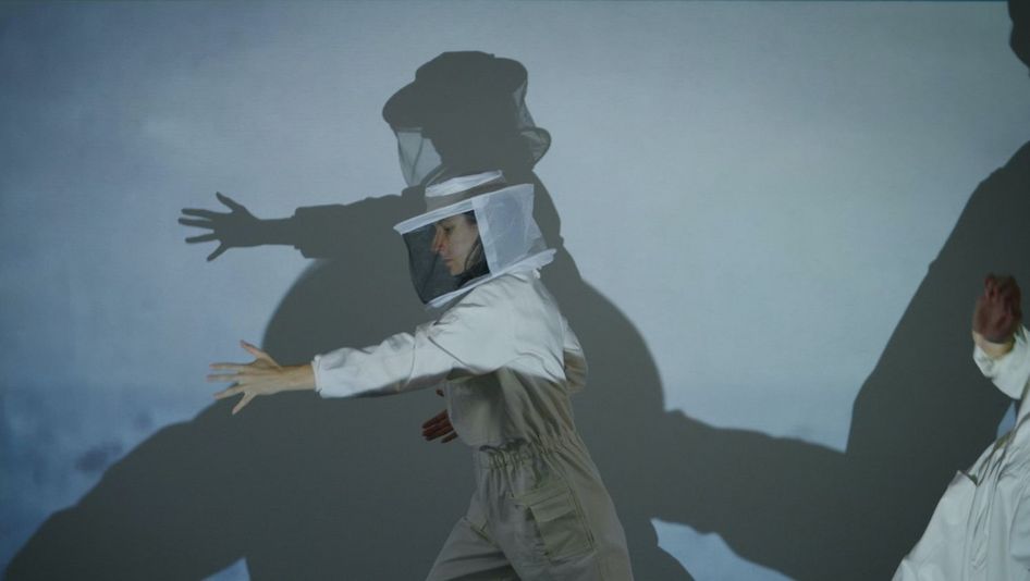 Filmstill aus WHERE GRAVITY FADES: Eine Person in einem weißen Imkeranzug performt etwas vor einer hellblauen Wand, wobei auch ihr Schatten an der Wand zu sehen ist.