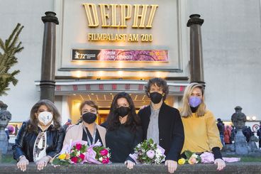 In front of the cinema, five people wearing masks and holding flowers are looking into the camera.