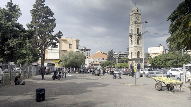 Stadtplatz mit Uhrturm, Marktstand und verstreuten Personen bei Tageslicht.