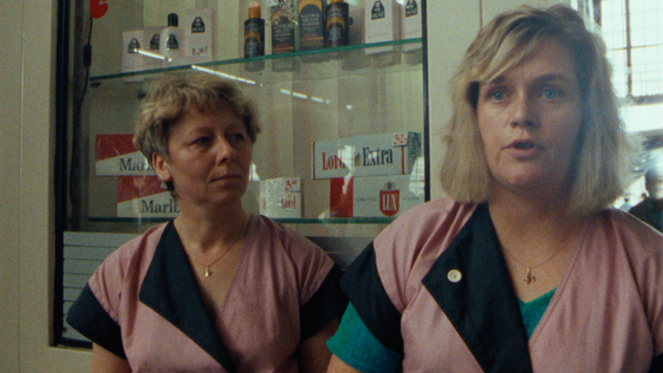 Filmstill aus BERLIN, BAHNHOF FRIEDRICHSTRASSE 1990: Zwei Verkäuferinnen in rosa-schwarzen Arbeitskitteln stehen vor einer Auslage mit Zigaretten und Kosmetikartikeln.