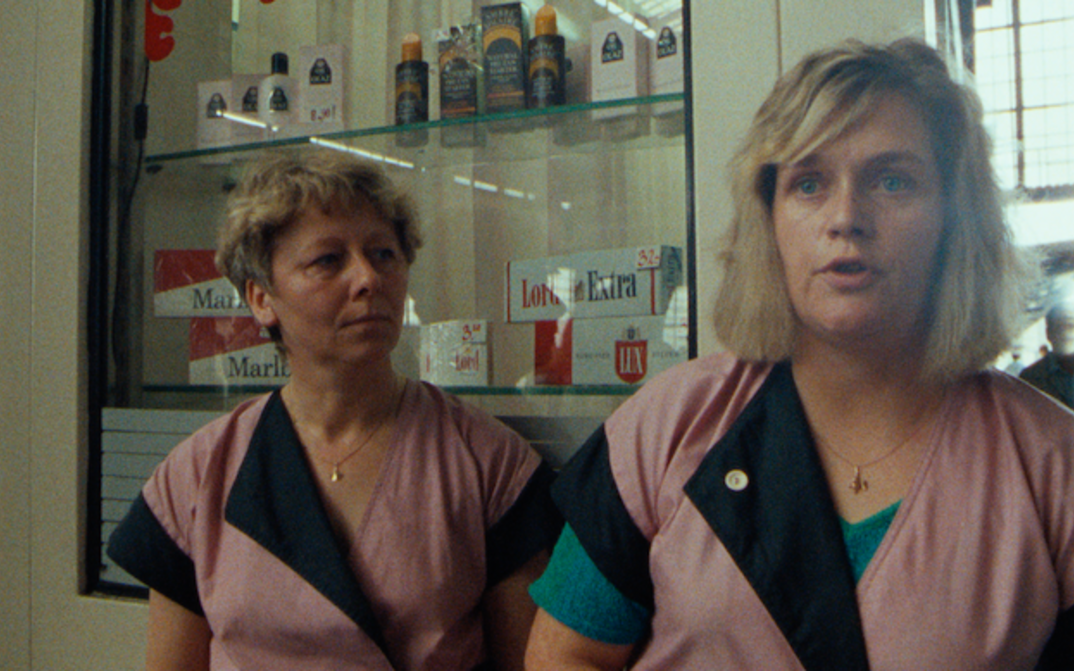 Filmstill aus BERLIN, BAHNHOF FRIEDRICHSTRASSE 1990: Zwei Verkäuferinnen in rosa-schwarzen Arbeitskitteln stehen vor einer Auslage mit Zigaretten und Kosmetikartikeln.