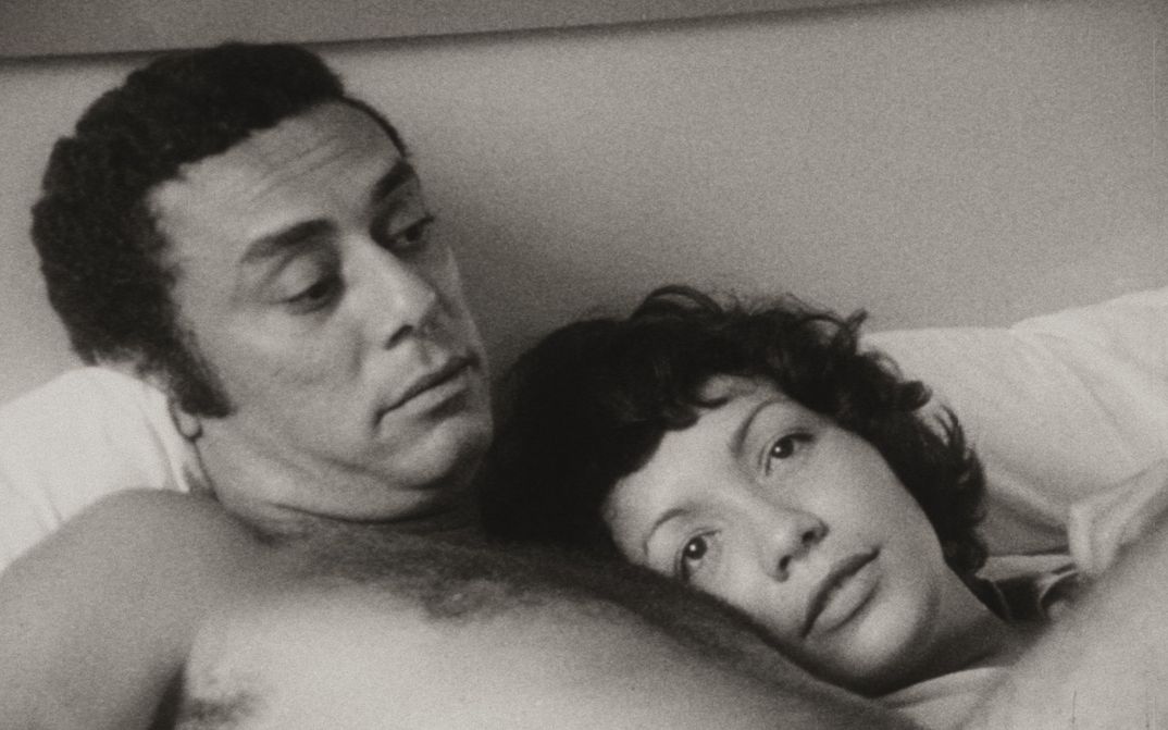 Film still from DE CIERTA MANERA: A man and a woman are lying in bed together. She has laid her head on his chest.