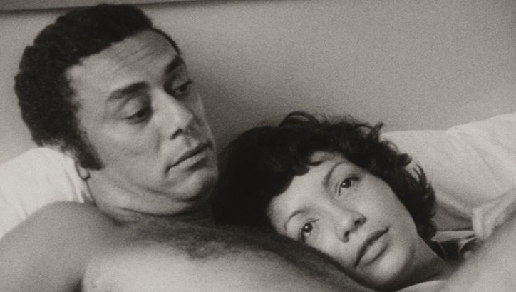 Film still from DE CIERTA MANERA: A man and a woman are lying in bed together. She has laid her head on his chest.
