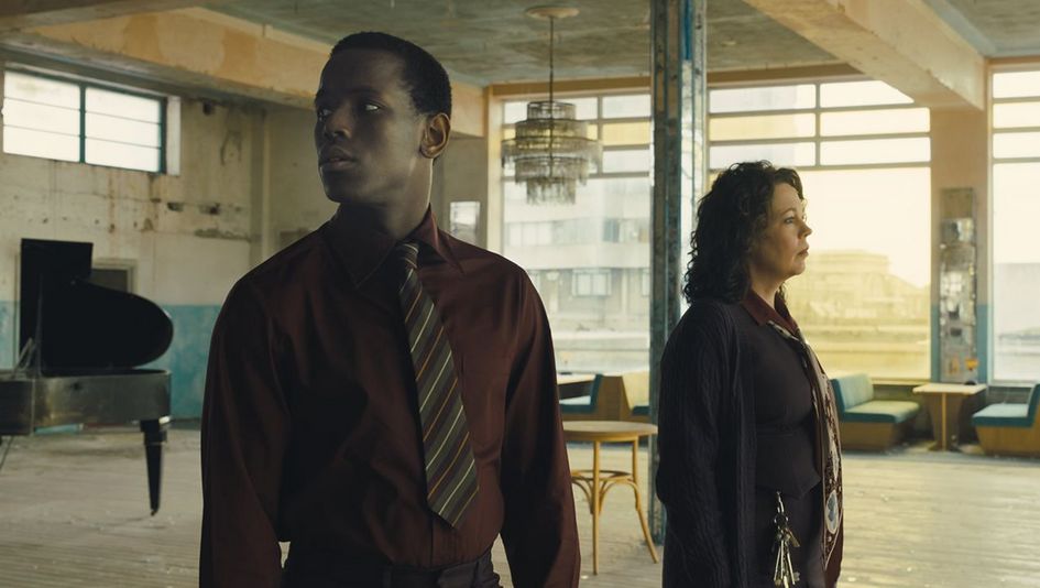 Film still from EMPIRE OF LIGHT: A man and a woman are standing in a large, seemingly deserted room and looking around. There are tables, benches, and a grand piano in the room.