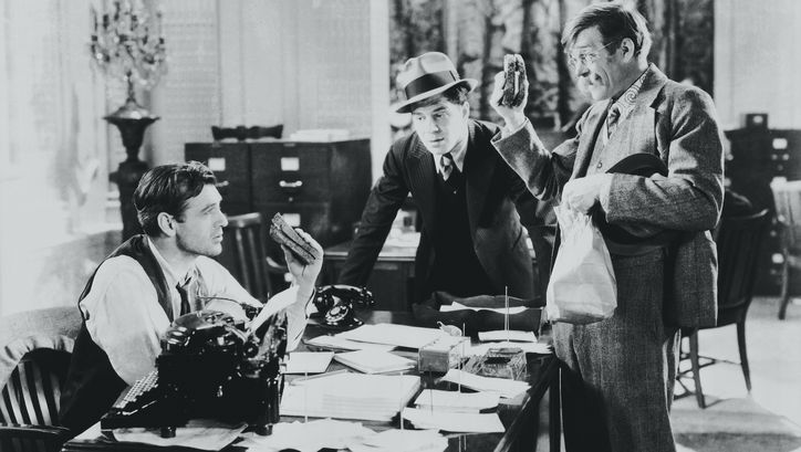 Film still from MR. DEEDS GOES TO TOWN: A journalist is sitting at a desk where two other men are standing. Two of the men are holding a sandwich.