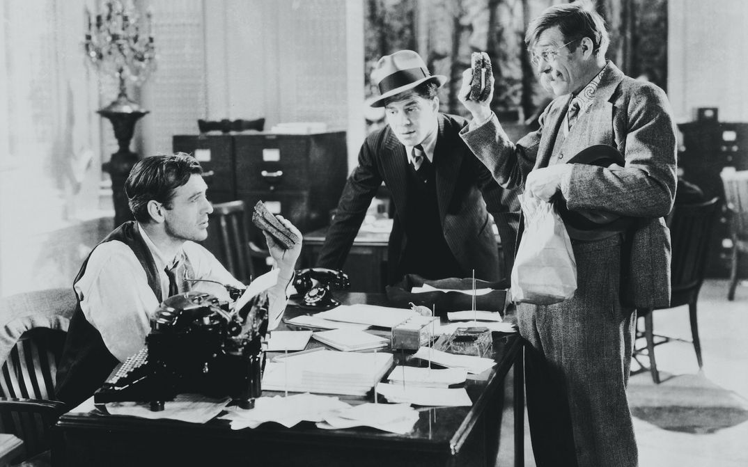 Film still from MR. DEEDS GOES TO TOWN: A journalist is sitting at a desk where two other men are standing. Two of the men are holding a sandwich.