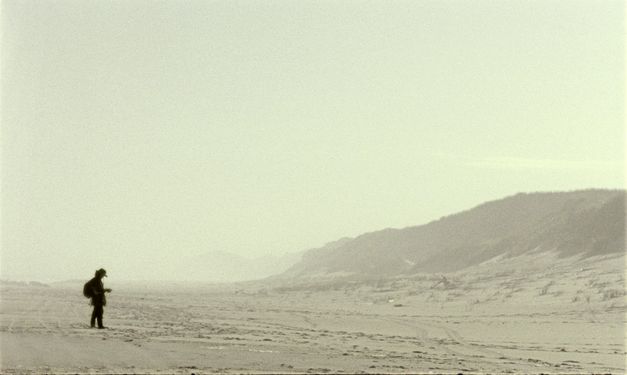 Still from the film "Geographies of Solitude" by Jacquelyn Mills. In a wide shot we see the small figure of a woman of a foggy beach. 