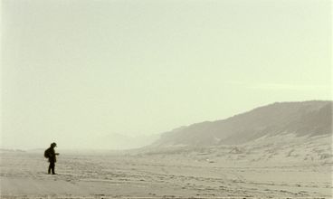 Filmstill aus „Geographies of Solitude“ von Jacquelyn Mills. Aus einiger Entfernung sehen wir eine Frau an einem leeren, nebligen Strand.