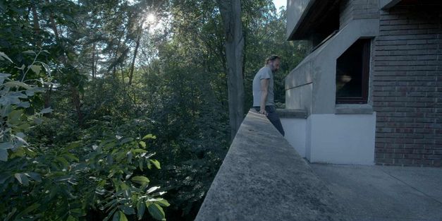Filmstill aus „We Haven’t Lost Our Way“ von Anka und Wilhelm Sasnal. Ein Mann lehnt an der Brüstung einer Terrasse. Im Hintergrund sehen wir grünen Wald.   