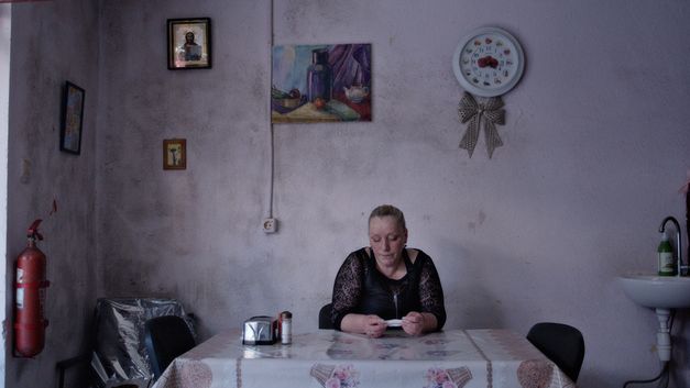 Film still from WHEN LIGHTNING FLASHES OVER THE SEA by Eva Neymann. The picture shows a woman sitting at a table with her head down. A clock and pictures hang on the wall.