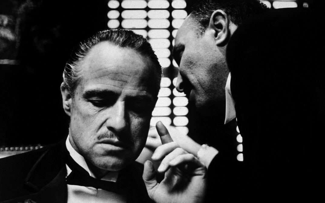 Filmstill aus THE GODFATHER: Ein Mann flüstert einem anderen etwas ins Ohr.