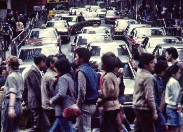 Film still from HONG KONG TOPOGRAPHY. People crossing a street, cars in the background.