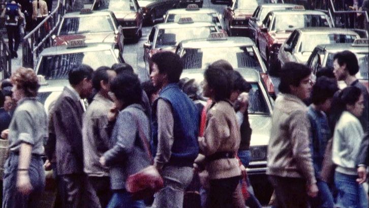 Filmstill aus HONG KONG TOPOGRAPHY. Menschen überqueren eine Straße, im Hintergrund Autos.