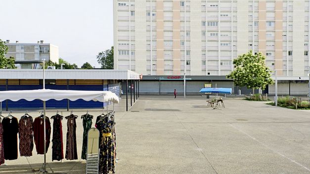 Filmstill aus "Europe" von Philipp Scheffner. Ein leerer Platz in einer Hochhaussiedlung. Im Vordergrund ein Stand mit Kleidung. 