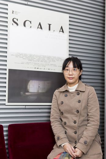 A woman sitting on a cinema chair in front of a wall is looking into the camera. On the wall there is a poster with the title “Scala“.
