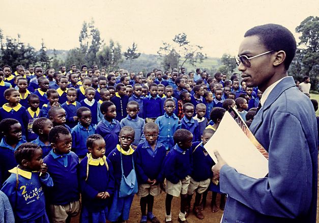 Still from the film „Der Kampf um den heiligen Baum" by Wanjiru Kinyanjui. It shows a large group of children standing in front of a man in a suit.