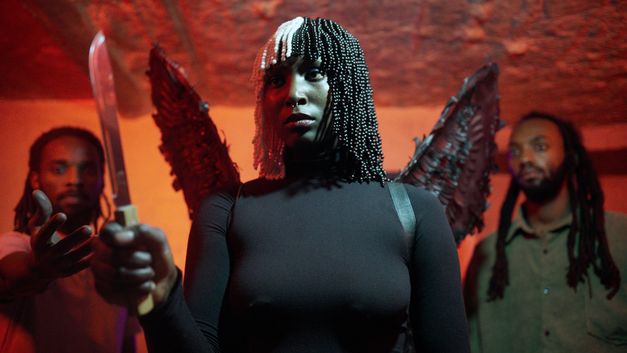 Film still from MINIMALS IN A TITANIC WORLD by Philbert Aimé Mbabazi Sharangabo. The picture shows a person dressed in black with wings and a knife in her hand. Two other people are standing behind her on the left and right.