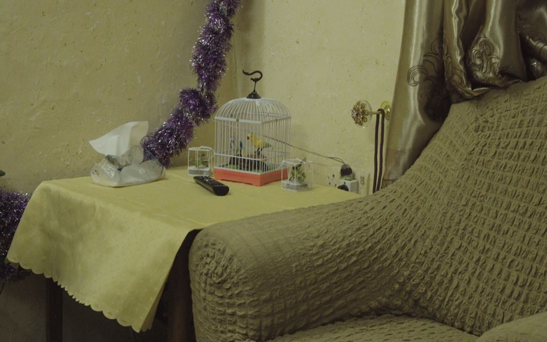 Still from “J-N-N” by Ginan Seidl. In a corner of a living room captured in a close shot, a beige armchair stands next to a wooden table. On it lie a yellow tablecloth, a remote control, and a decorative bird in a cage. A sparkly purple garland placed against the wall contrasts with the ensemble. 
