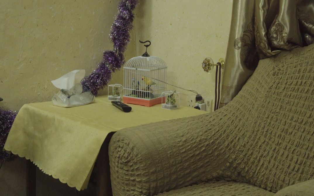 Filmstill aus dem Film „ J-N-N“ von Ginan Seidl. In einer in Nahaufnahme aufgenommenen Wohnzimmerecke steht ein beiger Sessel neben einem Holztisch. Darauf liegen eine gelbe Tischdecke, eine Fernbedienung und eine dekorative Vogelfigur in einem Käfig. Eine glitzernde violette Girlande an der Wand bildet einen Kontrast zum Ensemble.