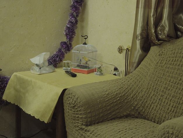 Still from “J-N-N” by Ginan Seidl. In a corner of a living room captured in a close shot, a beige armchair stands next to a wooden table. On it lie a yellow tablecloth, a remote control, and a decorative bird in a cage. A sparkly purple garland placed against the wall contrasts with the ensemble. 
