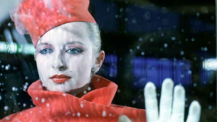 Film still from BILDNIS EINER TRINKERIN: An elegantly dressed and made-up woman stands behind a window pane on which raindrops can be seen.