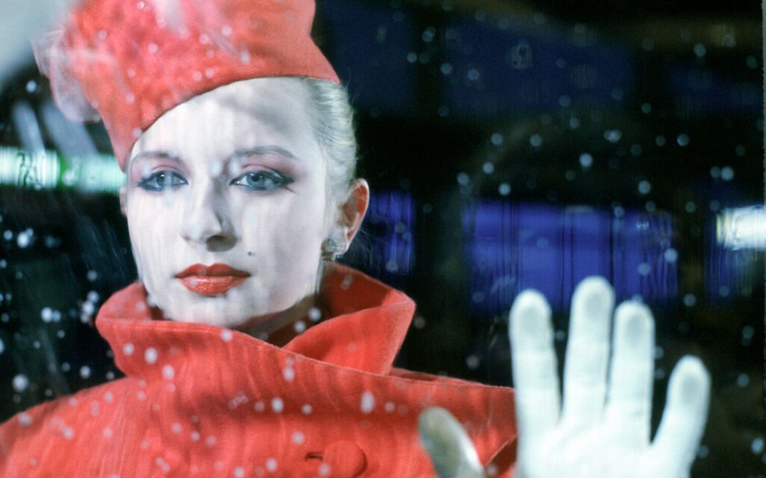 Filmstill aus BILDNIS EINER TRINKERIN: Eine elegant gekleidete und geschminkte Frau steht hinter einer Fensterscheibe, auf der Regentropfen zu sehen sind.