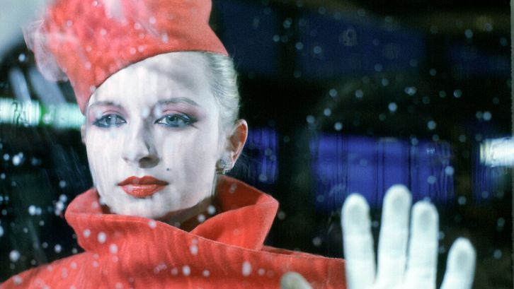 Filmstill aus BILDNIS EINER TRINKERIN: Eine elegant gekleidete und geschminkte Frau steht hinter einer Fensterscheibe, auf der Regentropfen zu sehen sind.