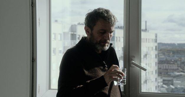 Still from the film "Le Gang des Bois du Temple" by Rabah Ameur-Zaïmeche. A man with grey hair, a grey beard and a brown sweater is standing in front of a window and looking down at a glass of water in his right hand. There is a multistory apartment complex in the background.