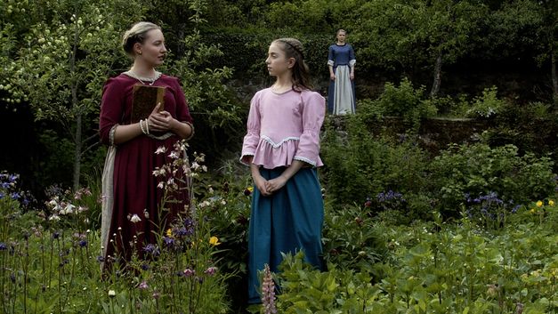 Filmstill aus "Ihre ergebenste Fräulein" von Eva C. Heldmann. Zu sehen sind drei Frauen in einem Garten. Zwei Frauen stehen im Vordergrund und unterhalten sich. Die Frau im Hintergrund schaut in ihre Richtung. Sie tragen häusliche Kleidung aus dem 18. Jahrhundert. 