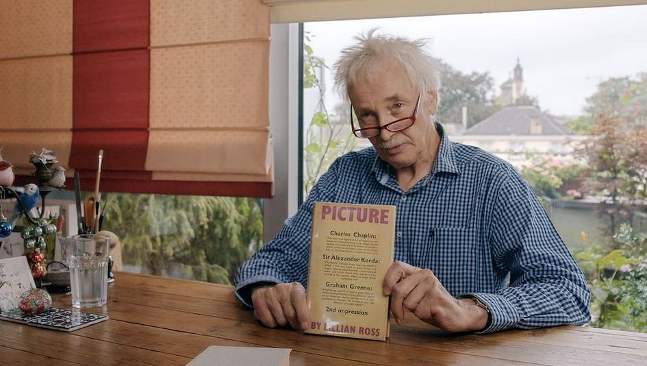 Filmstill aus ERZÄHLUNGEN EINES KINOGEHERS – WERNER DÜTSCH: Ein älterer Mann (Werner Dütsch) mit weißen strubbeligen Haaren sitzt an einem Tisch vor einem großen Fenster und hält ein Buch in die Kamera.